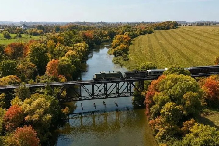 Train crossing a bridge over a river with autumn-colored trees and a field.