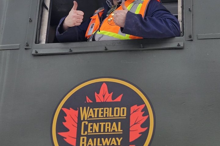 Person with a green hat and safety vest gives thumbs up from a railway train window.