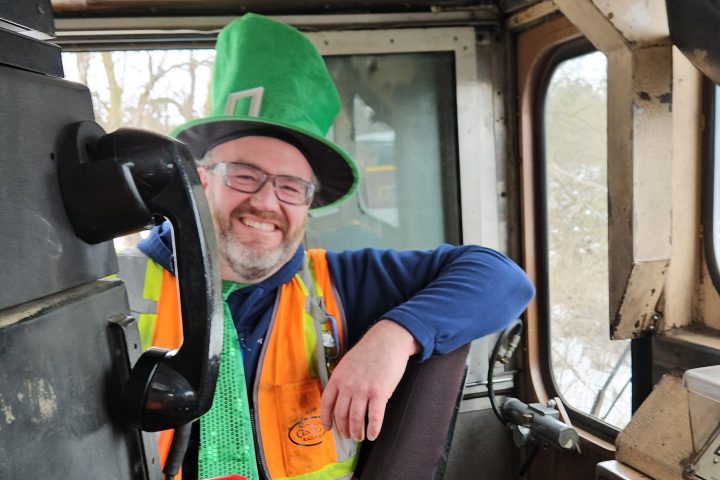 Man in a green hat and safety vest smiles in a train cab.