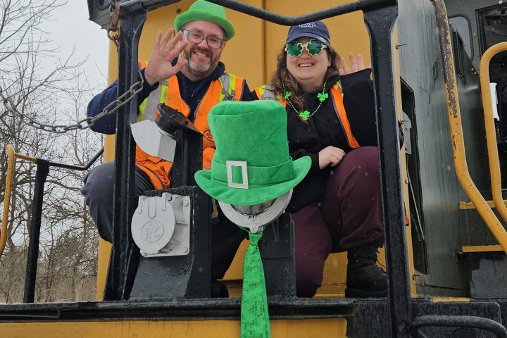 Two people in festive green hats wave from a train numbered 1012 with a leprechaun hat on the front.