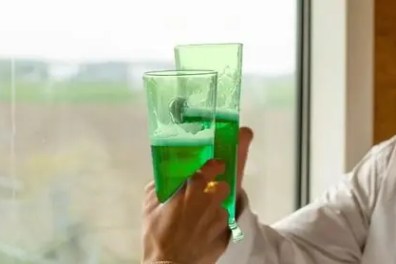 Two people toasting with green drinks by a window.