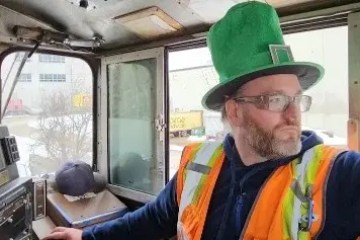 Person in a train cab wearing a green hat and orange safety vest, looking out of the window.