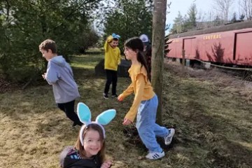 Children outdoors hunting for eggs near a train, one wearing blue bunny ears.