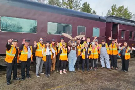 Group of people in orange vests pose and wave beside a maroon train.