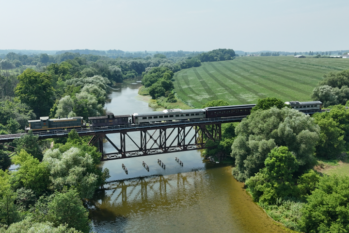 Train crosses a metal bridge over a river surrounded by lush green fields and trees.