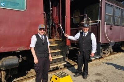 Two uniformed train conductors standing by a vintage maroon train car.