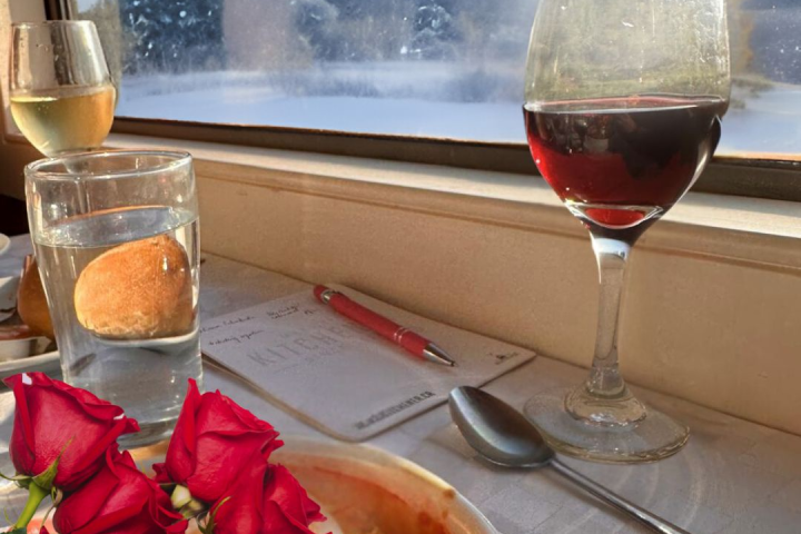 Table with red wine, glass of water, roses, and snowy view through window.