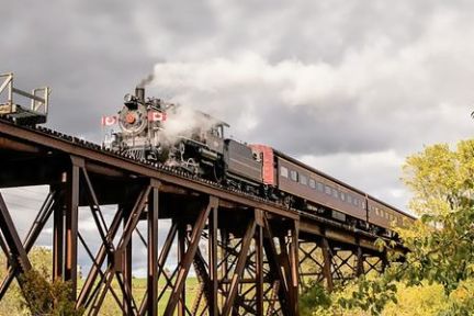 Steam train crossing a tall metal bridge with cloudy sky background.