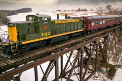 Green and yellow train crossing a snowy bridge in a rural landscape.