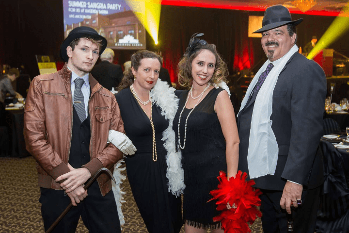 Four people in 1920s attire posing at an indoor event with dim lighting.