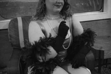 Person in vintage attire sitting with gloves and pearls in a train carriage.