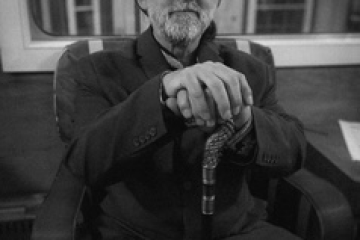 Elderly man with hat and cane sitting indoors, black and white photo.