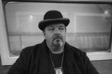 Man in bowler hat and coat, wearing a badge, sits in a room with a window, black and white photo.