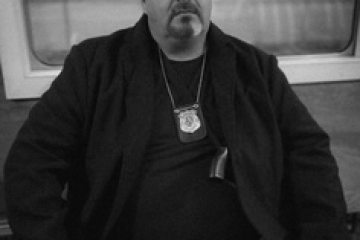 Man in bowler hat and coat, wearing a badge, sits in a room with a window, black and white photo.
