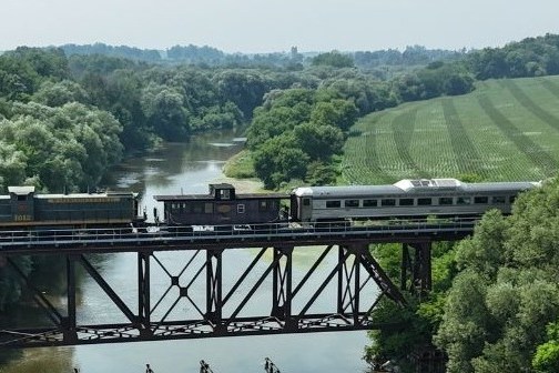 Train on a bridge over a river with trees and fields in the background.