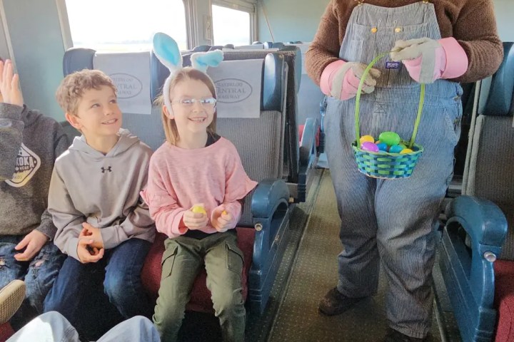 Children on train smiling at person in Easter Bunny costume holding a basket of colorful eggs.
