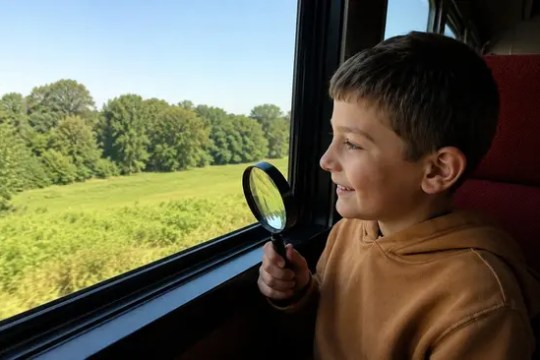 Child with magnifying glass looking out the train window at green landscape.