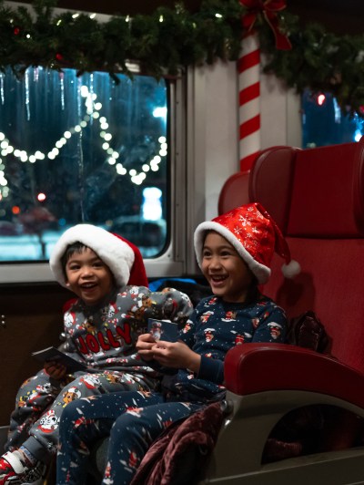 Two children in Christmas pajamas and Santa hats laugh aboard a decorated train.