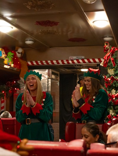 Two people in elf costumes entertaining passengers on a festive train decorated with Christmas ornaments.