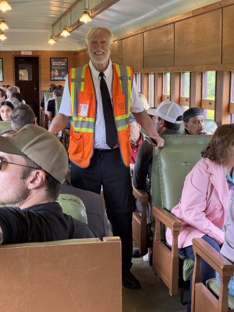 Train conductor in orange vest talks to seated passengers inside a train carriage.
