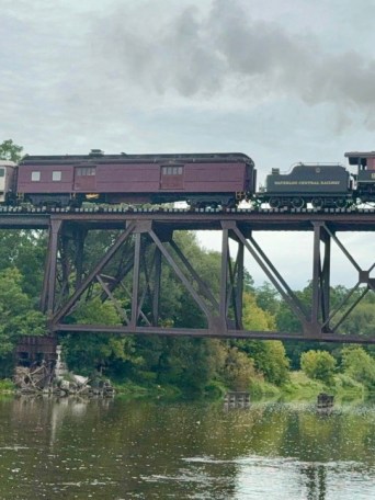 Steam train crossing a tall bridge over a river amid lush greenery.