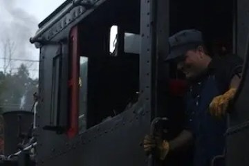 Person operating a vintage steam locomotive, wearing a cap and gloves.