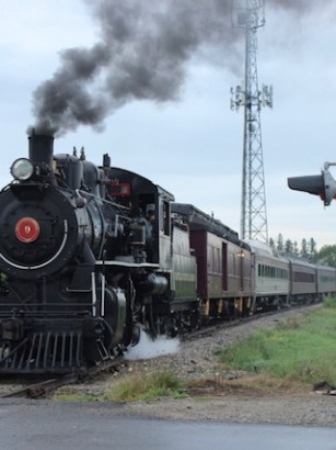 Steam train crossing a rural railroad crossing on a cloudy day.