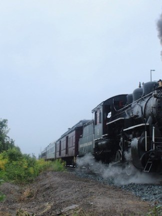 Steam locomotive on tracks with smoke billowing in a countryside setting.
